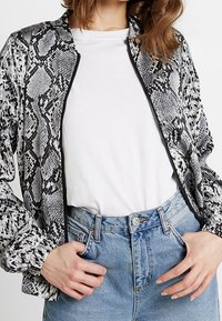 Chaqueta bomber con estampado de piel de serpiente en blanco y negro, con cierre de cremallera, tela suave y puños elásticos, combinada con una camiseta blanca y jeans de color azul claro.