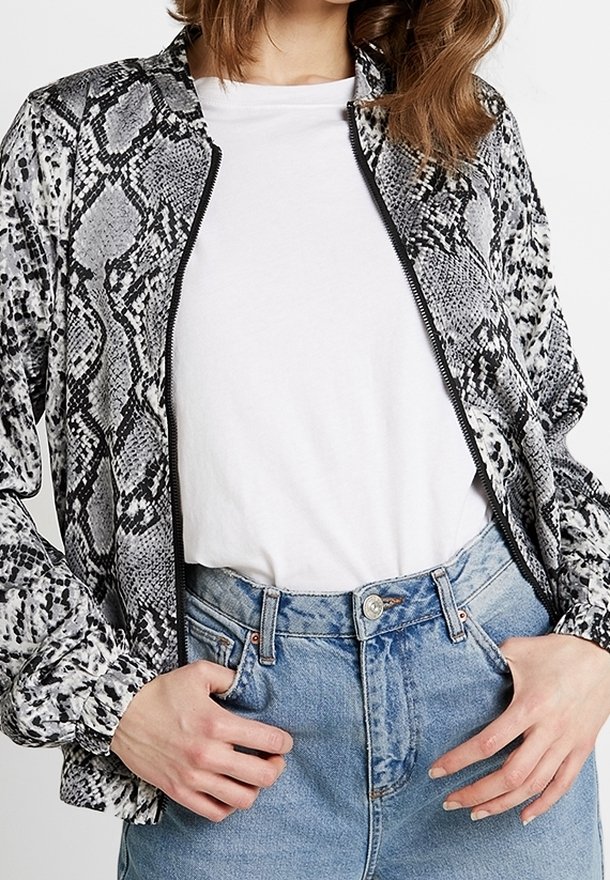 Chaqueta bomber con estampado de piel de serpiente en blanco y negro, con cierre de cremallera, tela suave y puños elásticos, combinada con una camiseta blanca y jeans de color azul claro.