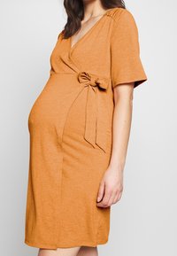 Czerwono-pomarańczowa sukienka ciążowa z dekoltem w kształcie V, krótkimi rękawami, detalem w postaci wiązania z boku oraz dopasowanym krojem podkreślającym ciążowy brzuch.