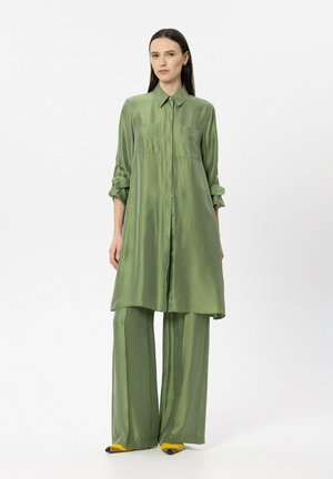 Frau trägt ein locker sitzendes, glänzendes olivgrünes langes Hemd mit hochgekrempelten Ärmeln und passende weit geschnittene Hosen, steht vor einem schlichten weißen Hintergrund.