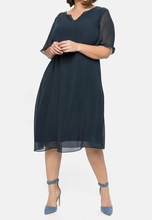 Mujer vestida con un vestido azul marino hasta la rodilla con mangas cortas transparentes y tacones altos azules con tiras en el tobillo, de pie frente a un fondo liso.