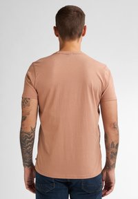 Tílko s krátkým rukávem, přiléhavého střihu v barvě jemné růžové, vyrobené z měkké tkaniny, s kulatým výstřihem a hladkým povrchem. Tetování viditelné na pažích.