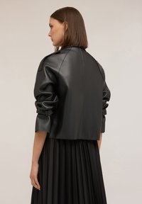 Giacca cropped in pelle nera con spalle arrotondate, maniche arricciate e dettagli di cucitura visibili, abbinata a una gonna nera plissettata.