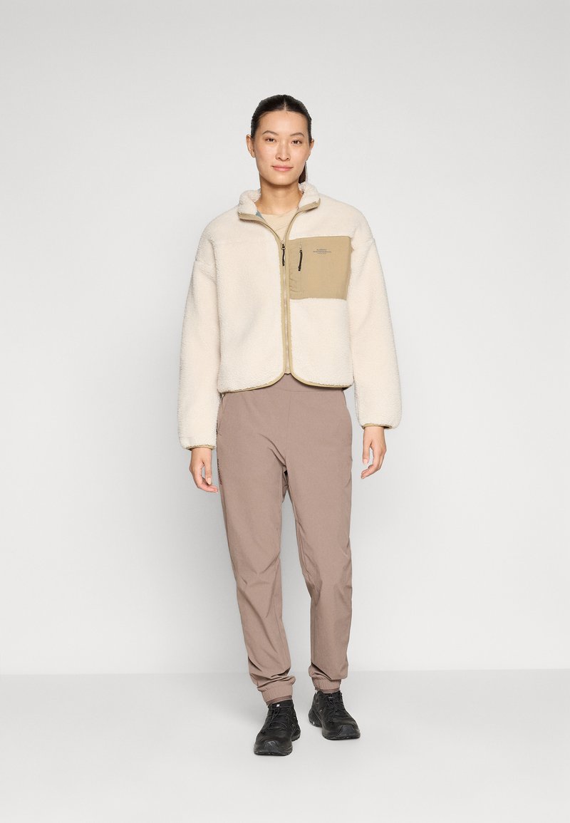 Giacca di pile crema con una tasca sul petto color sabbia, chiusura con zip e polsini elasticizzati, abbinata a pantaloni beige chiaro e scarpe nere.