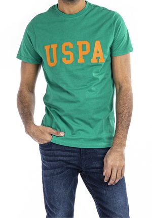 Mann trägt ein grünes T-Shirt mit kurzen Ärmeln und orangefarbenen "USPA" Buchstaben sowie dunkelblaue Jeans, eine Hand in der Tasche, vor weißem Hintergrund.
