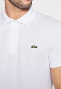 Polo shirt blanc en tissu texturé, arborant un logo de crocodile vert sur la poitrine gauche, deux boutons au col et des manches courtes.