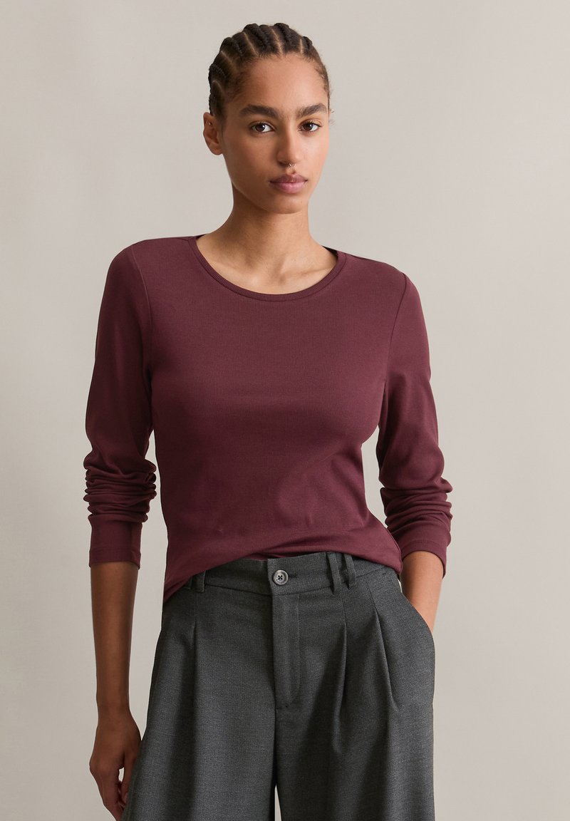 Burgunderfarget langermet topp med rund hals, kombinert med høytlivede grå plisserte bukser. Myk stoff med en tettsittende design.