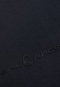 Svart strukturerad tyg med präglad "Sail Racing"-logotyp. Den släta ytan och subtila ribbade mönstret förbättrar materialets synlighet.