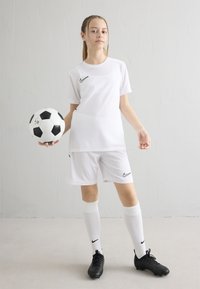 Nuori naisjalkapalloilija valkoisessa peliasussa, joka pitää mustavalkoista jalkapalloa kädessään ja seisoo yksiväristä vaaleaa taustaa vasten.