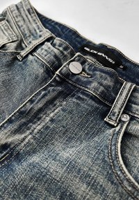 Denim jeans in een medium wash hebben een traditionele vijfzakkenontwerp, een zilveren metalen knoop en contrasterende stiksels. Etiket zichtbaar op de tailleband.