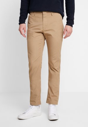 Mann trägt beige Chinos, weiße Sneakers und ein marineblaues Langarmhemd vor hellem, einfarbigem Hintergrund.