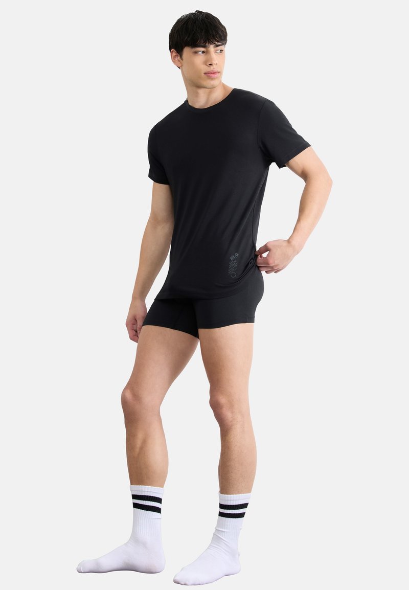 T-shirt noir à manches courtes et boxer noir à la coupe ajustée. Chaussettes blanches avec des rayures noires. Texture lisse, branding minimal.
