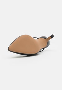 Black high-heeled shoe with a pointed toe and thin ankle strap. The sole is tan with a textured surface and a triangular shape.