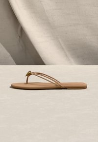 Μπεζ επίπεδη σάνδαλα με λεπτούς διπλούς ιμάντες και ένα μικρό χρυσό διακοσμητικό κουμπί, παρουσιάζεται σε φωτεινή επιφάνεια με φόντο μπεζ ύφασμα.