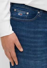 Jeans in denim blu scuro con un bottone in metallo argentato, logo ricamato sulla tasca posteriore e una superficie testurizzata. Mano posata sull'anca.