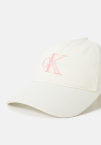 Boné em tom de creme com aba curva, apresentando o logótipo bordado "CK" em rosa pastel e branco na frente. Textura de tecido leve.