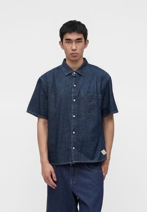 Ung mand med kort sort hår iført en mørkeblå kortærmet denimskjorte med knapper og matchende denimbukser, stående mod en hvid baggrund.
