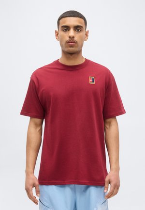Giovane uomo con capelli corti che indossa una maglietta rossa tinta unita con un piccolo logo multicolore sul petto e pantaloncini azzurri, in piedi davanti a uno sfondo bianco.