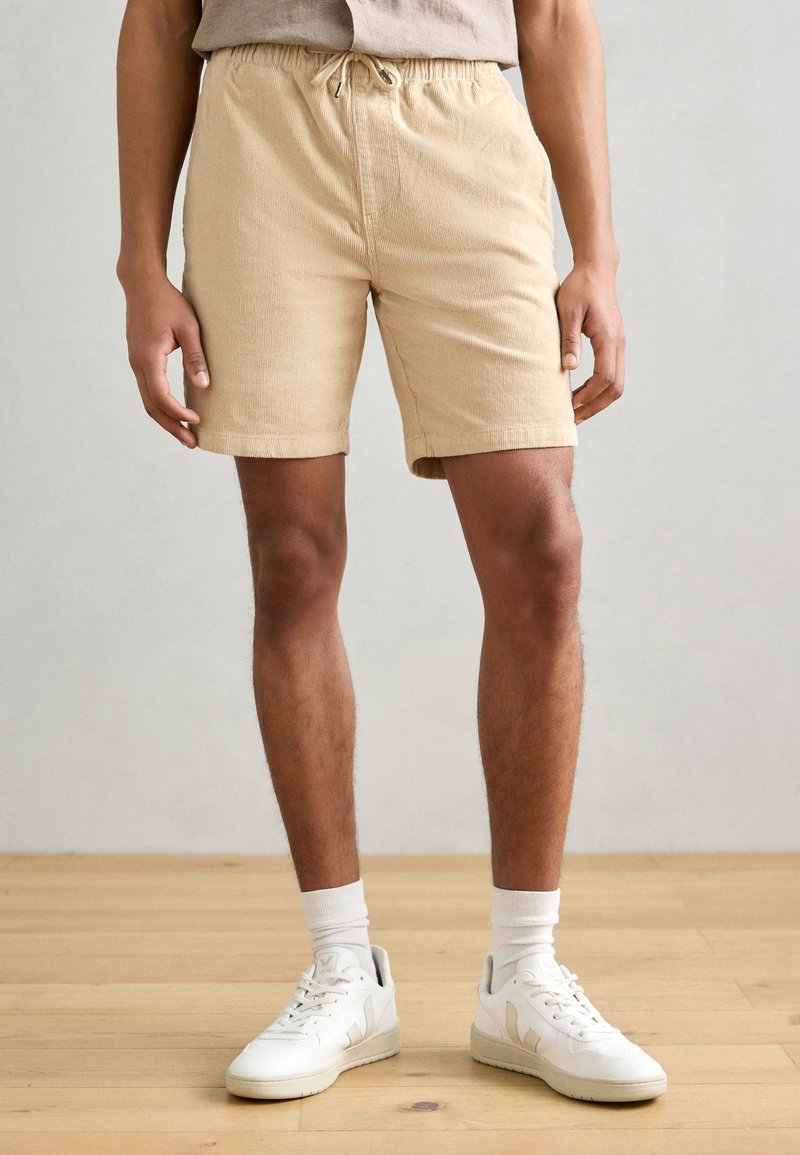 Shorts beiges texturés avec une taille élastique et un cordon de serrage. Associés à des baskets blanches et des chaussettes, présentés sur un fond neutre.
