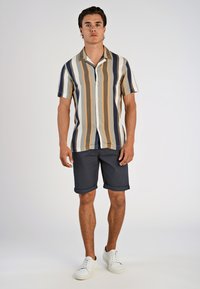 Kortermet skjorte med knapper og vertikale striper i beige, marineblå og sennep, kombinert med mørke rullede shorts og hvite joggesko.