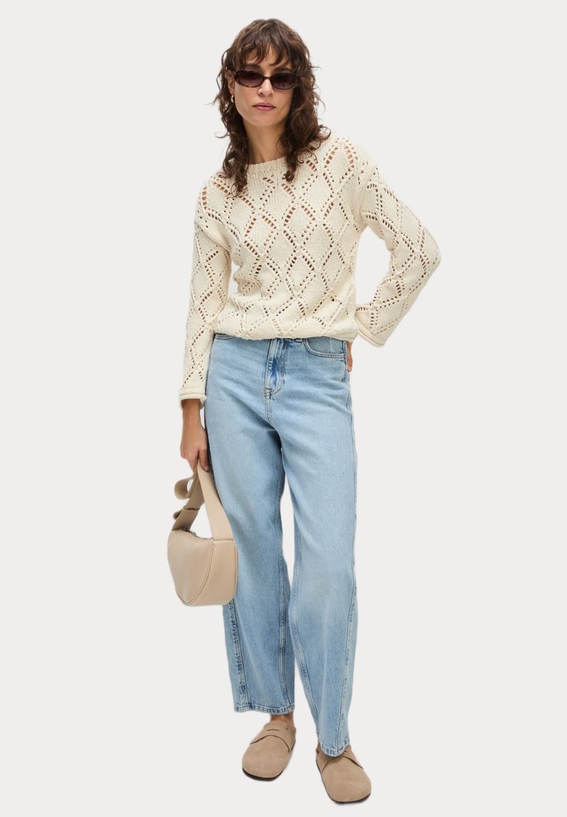 Crèmekleurige gebreide trui met ruitpatroon, gecombineerd met lichtblauwe hoge taille jeans en beige sandalen. Houdt een beige handtas vast.