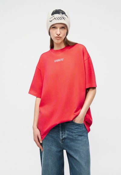 Γυναίκα που φοράει μια κόκκινη φαρδιά μπλούζα με το κείμενο «STIEGLITZ», μπλε τζιν και άσπρο σκούφο με μαύρο σχέδιο προσώπου, πόζα με το ένα χέρι στην τσέπη.