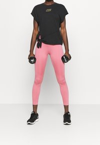 Camiseta negra de Nike con detalle de lazo; leggins rosas; zapatillas negras. Persona sosteniendo mancuernas, de pie en una posición neutral.