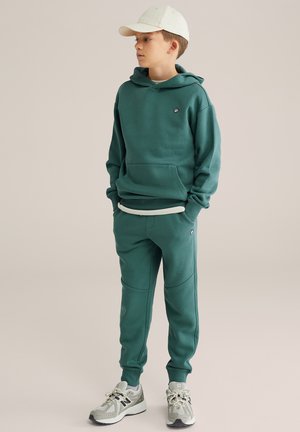 Zelená mikina s kapucňou a predným vreckom, skombinovaná so zodpovedajúcimi teplákmi s bočnými panelmi. Doplnkom je béžová čiapka a sivé bežecké topánky.