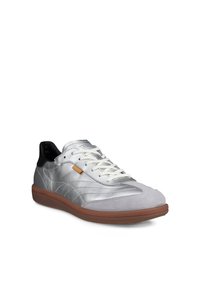 Baskets basses argentées avec lacets blancs, col de talon noir, languette décorative beige et semelle en caoutchouc marron, positionnées légèrement en angle.