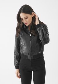 Giacca bomber nera in pelle con polsini e orlo a coste. Presenta due tasche frontali con zip e una finitura liscia. Il modello indossa pantaloni neri.