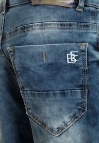 Jeans in denim con una lavatura blu sbiadita, con una tasca posteriore dotata di un logo ricamato bianco e dettagli di cucitura color marrone.