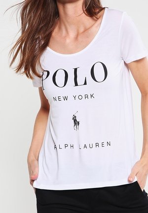 Vrouw draagt een wit T-shirt met zwarte tekst "POLO NEW YORK ALPH LAUREN" en een klein polo speler logo in het midden.