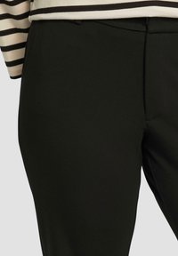 Pantalons noirs ajustés en tissu lisse, présentant un devant plat et des poches latérales, associés à une chemise à rayures partiellement visible au-dessus.
