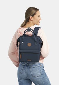 Marineblauwe stofrugzak met dubbele handgrepen en schouderbanden, voorzien van een voorzak met rits, logopatch en subtiele hardware accenten.