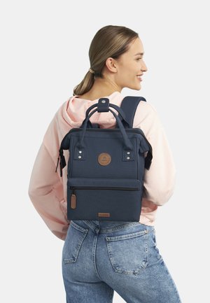 Sac à dos en tissu bleu marine avec doubles poignées et bretelles, comprenant une poche avant zippée, un patch logo et des accents métalliques discrets.
