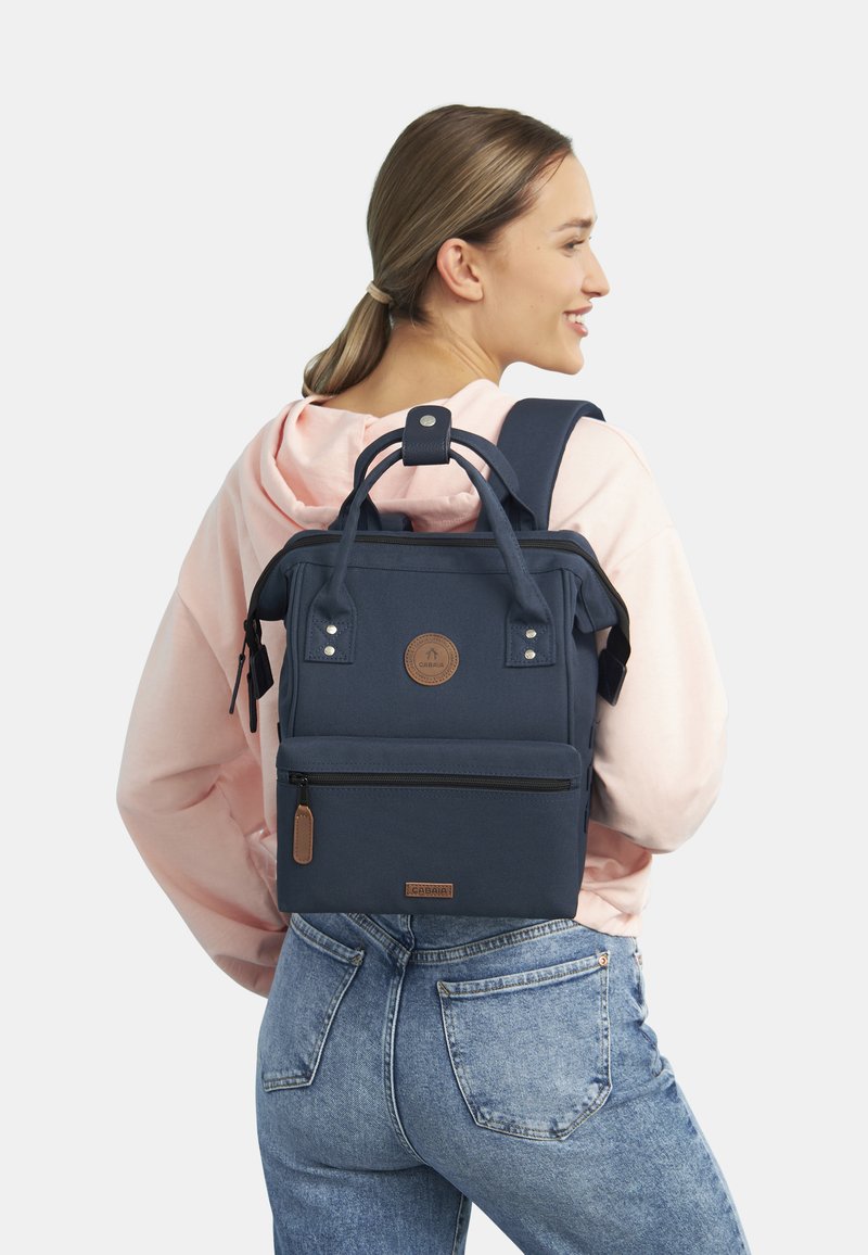 Marineblauwe stofrugzak met dubbele handgrepen en schouderbanden, voorzien van een voorzak met rits, logopatch en subtiele hardware accenten.