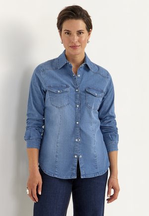 Vrouw met kort bruin haar draagt een blauw denim overhemd met knopen en donkere spijkerbroek, staand tegen een effen witte achtergrond.