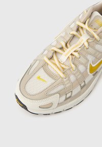 Μπεζ αθλητικό παπούτσι με επάνω μέρος από δίχτυ, κίτρινα στοιχεία και γκρι λεπτομέρειες. Διαθέτει σύστημα κορδονιών και λαστιχένια εξωτερική σόλα για πρόσφυση.