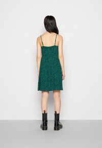 Mujer vestida con un vestido sin mangas de estampado de leopardo verde y tirantes finos, con botas negras hasta el tobillo, de pie frente a una pared blanca lisa.