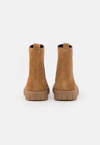 Brązowe zamszowe botki za kostkę z żebrowanymi gumowymi podeszwami, z elastycznymi bocznymi panelami i uchwytami do łatwego zakładania. Gładka tekstura z minimalnymi akcentami.
