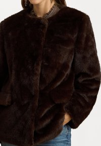 Bézs színű műszőrme kabát kerek nyakkivágással, gombos záródással és oldalsó zsebekkel. A puha textúra és a dús megjelenés fokozza a meleget és a kényelmet.
