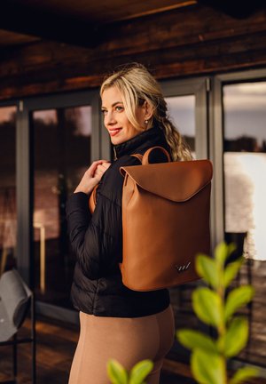 Tan leather backpack with a flap closure and logo detail, accompanied by a black puffer jacket and high-waisted beige leggings.