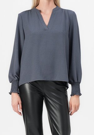 Kobieta ubrana w ciemnoszarą bluzkę z długim rękawem, ze ściągaczami przy mankietach i dekoltem w kształcie litery V, w połączeniu z czarnymi skórzanymi spodniami.