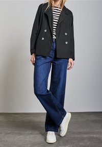 Persona vestita con un blazer doppiopetto nero, camicia a righe, jeans larghi blu e sneakers bianche, in piedi con le gambe incrociate.