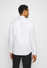 Camisa blanca de manga larga con corte recto y cuello estrecho, con puños de botones y una textura de tela suave. Se muestra la vista trasera.