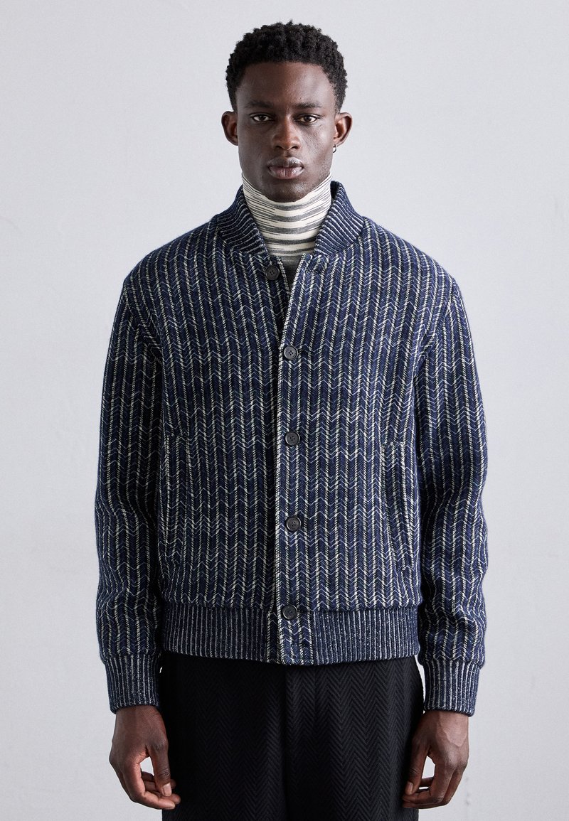 Mornarsko pleteno jopico z valovitim vzorcem, ki ima šal ovratnik, zapiranje na gumb in rebraste manšete. Nosi se čez črtast turtleneck.