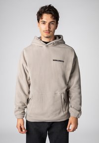 Beige Fleece-Kapuzensweatshirt mit einer Fronttasche, schwarzem "WORLDWIDE"-Text, Kordelzugkapuze und elastischen Bündchen vor hellgrauem Hintergrund.