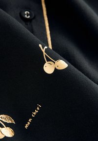 Černá tkanina s hladkým povrchem, zdobená zlatou grafikou třešní a textem "mon chéri" v kontrastním písmu. Viditelný detail knoflíku.