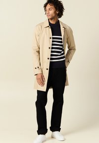 Trench coat beige con bottoni neri, indossato sopra un maglione a righe navy, abbinato a pantaloni neri e sneakers bianche.