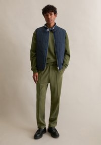 Marineblau gestepptes Weste über einem olivgrünen Sweatshirt und passenden Hosen. Schwarze Schuhe mit glatter Oberfläche. Neutraler Hintergrund.